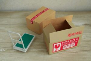 Open cardboard box with a red 'Fragile' label on a wooden floor, beside a small white-and-green hand truck or dolly nearby.