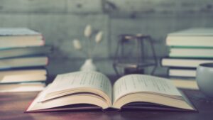 Open book laid flat on a wooden desk with stacked books, a lantern, and a cup in a cozy study setting