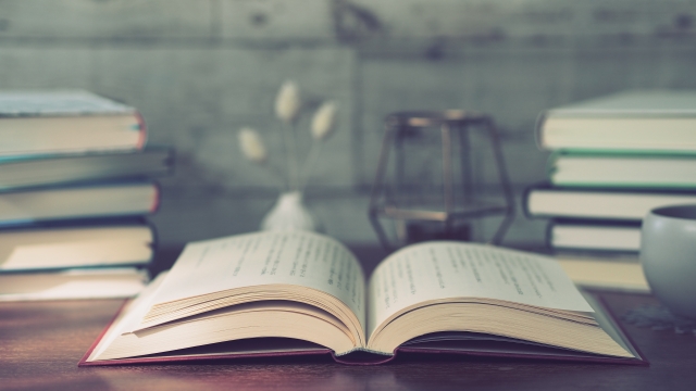 Open book laid flat on a wooden desk with stacked books, a lantern, and a cup in a cozy study setting