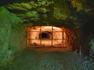 Underground cave tunnel with a metal barred gate blocking the passage and warm orange light beyond.