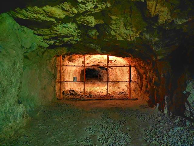 Underground cave tunnel with a metal barred gate blocking the passage and warm orange light beyond.
